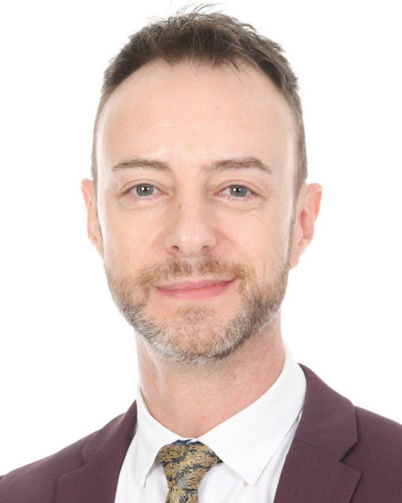 Professional headshot of a smiling man with light stubble, wearing a burgundy suit and patterned tie against a white background.